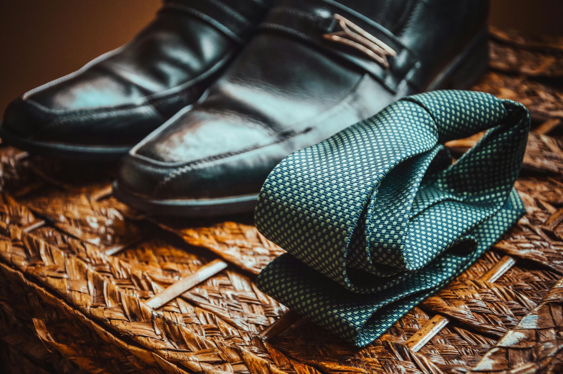 Black dress shoes and a rolled-up teal tie on a brown wicker surface.