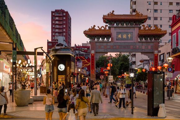 Calle peatonal muy concurrida al anochecer, con un arco tradicional chino, faroles y tiendas iluminadas.