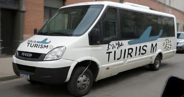 Minibús blanco con el logotipo de “Turismo” estacionado en una calle, vista lateral.