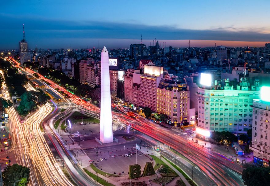 Vista nocturna de un bulevar de la ciudad con un obelisco iluminado y coloridas estelas de tráfico.