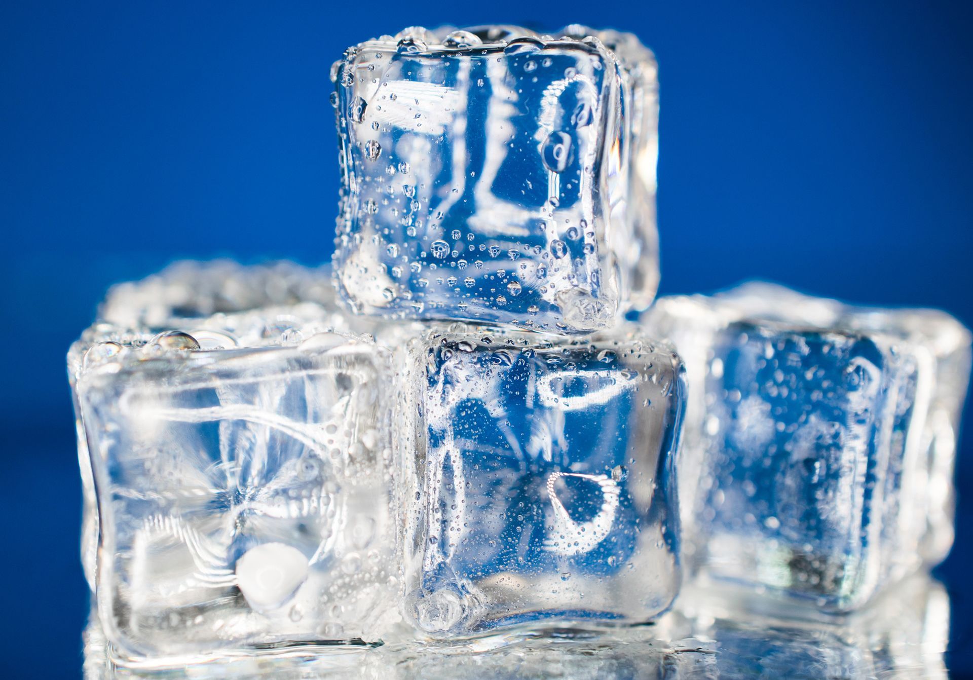 Close-up of transparent cubed ice stacked on reflective surface with blue background.
