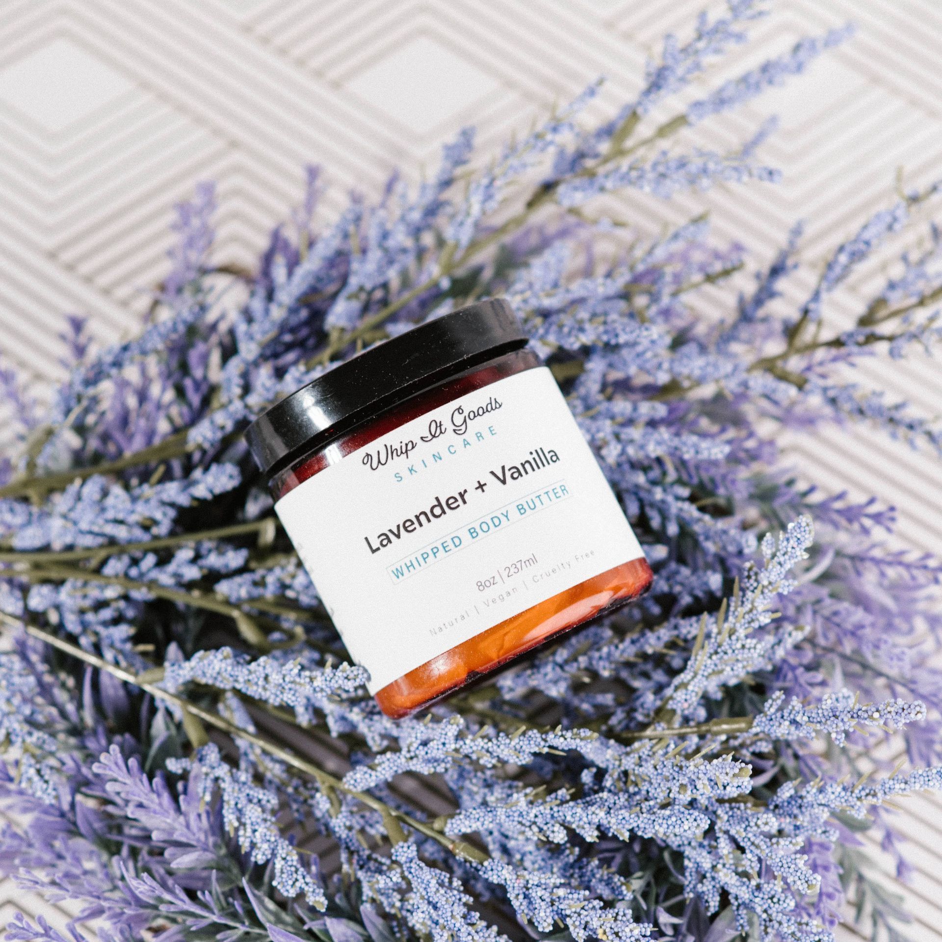 A jar of lavender and vanilla body butter is sitting on top of a bunch of lavender flowers.