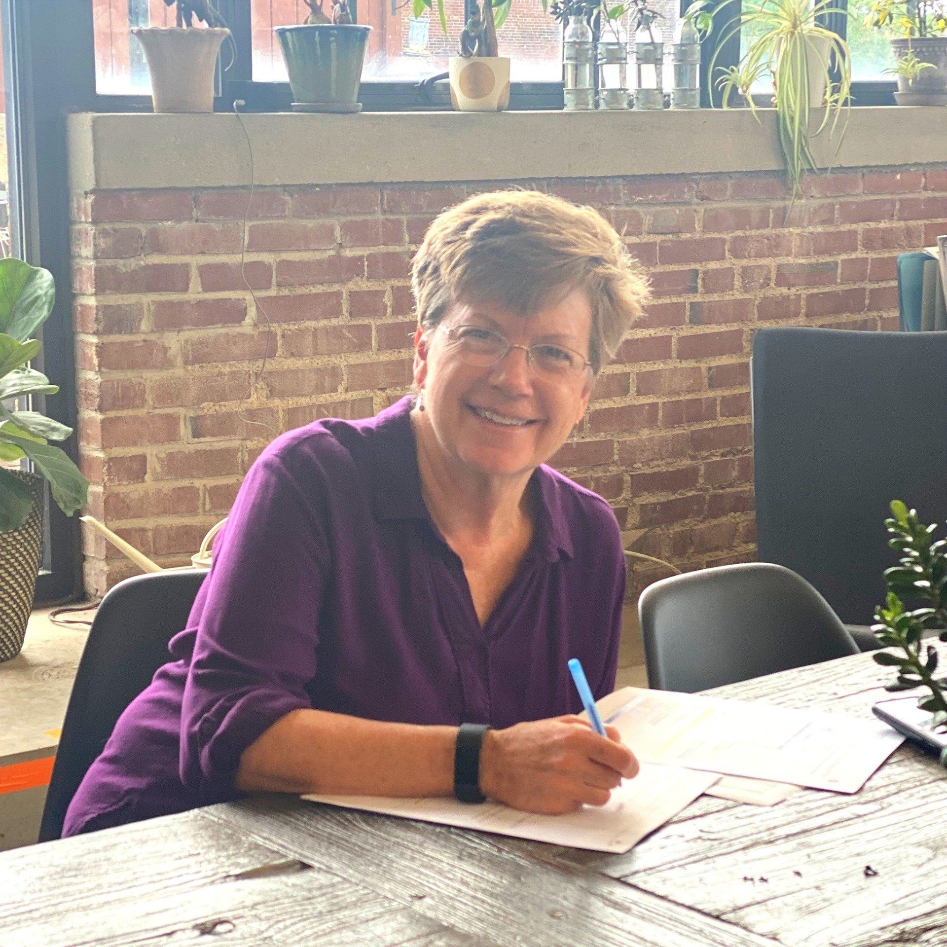 A woman in a purple shirt is sitting at a table writing on a piece of paper.