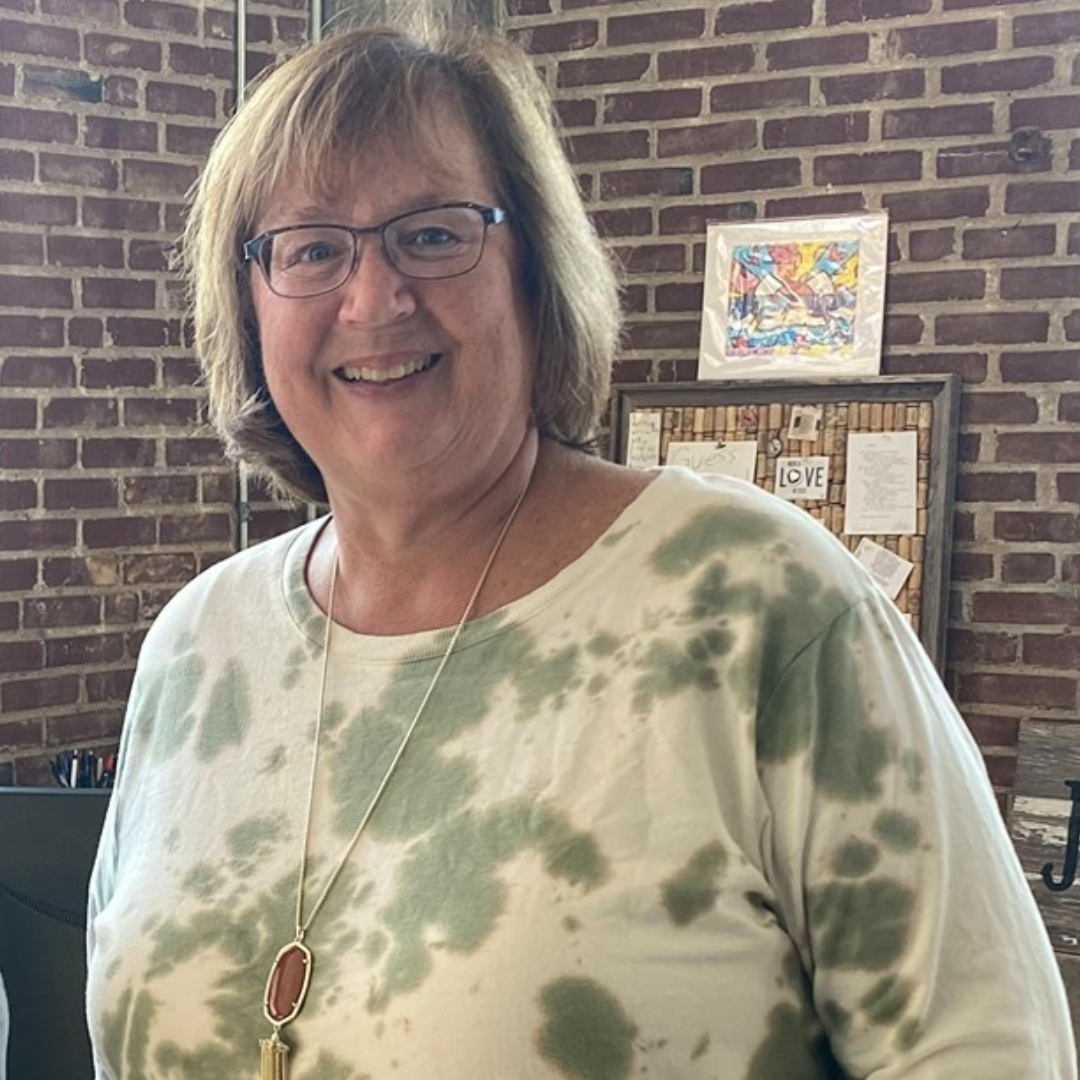 A woman wearing glasses and a tie dye shirt is smiling in front of a brick wall.