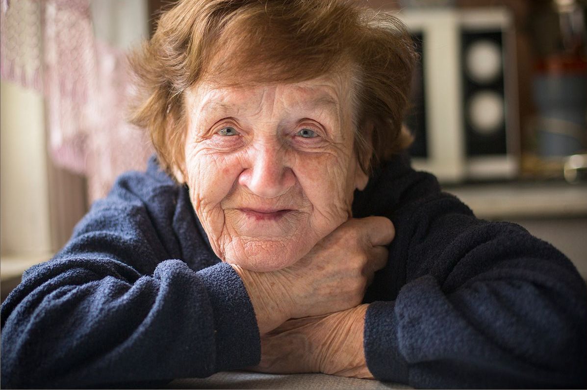 An elderly woman is sitting at a table with her hands on her chin.