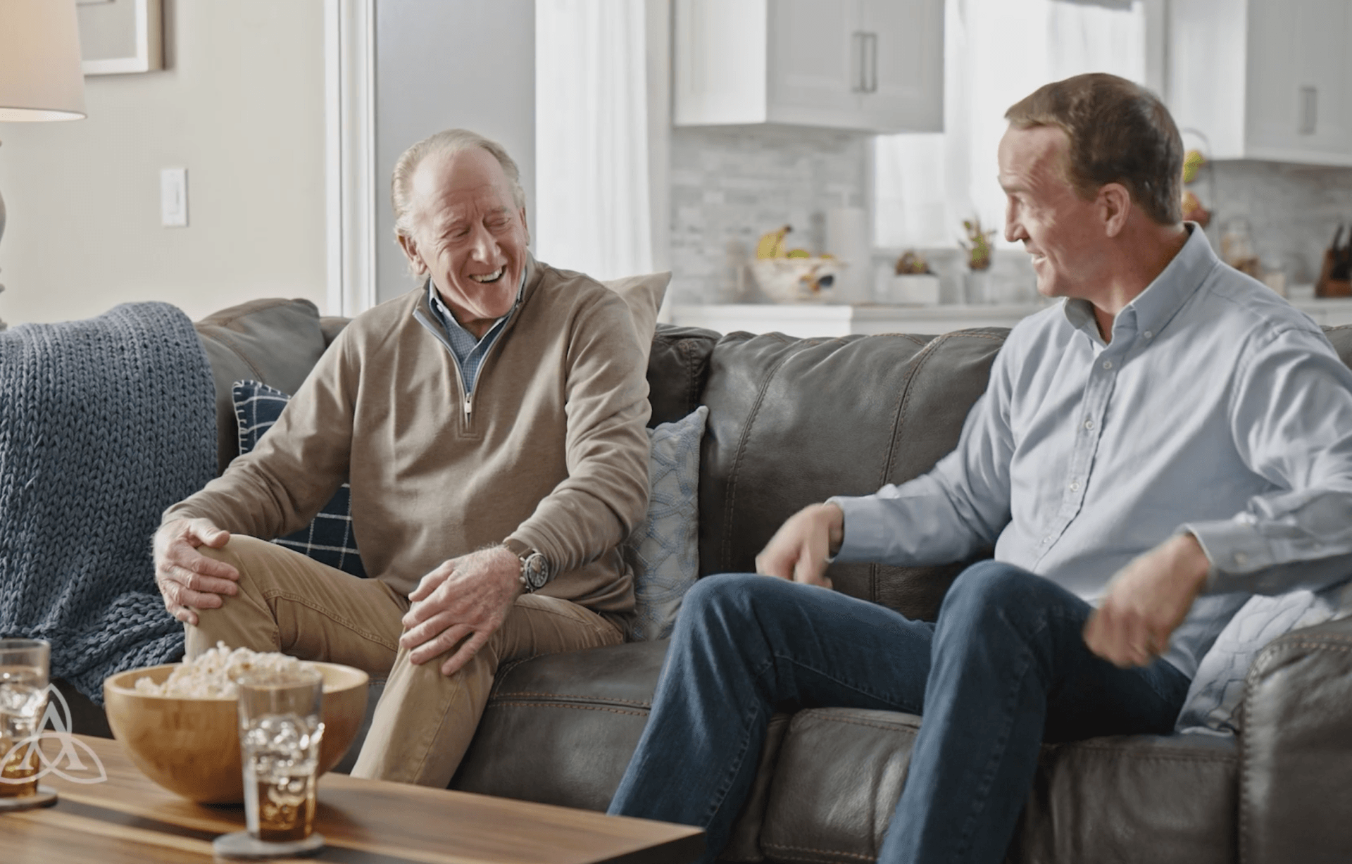 Peyton Manning and Archie Manning are sitting on a couch talking to each other.