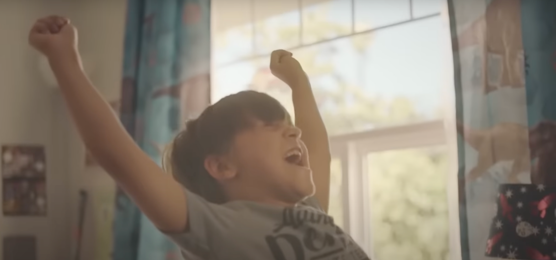 A young boy is standing in front of a window with his arms in the air.