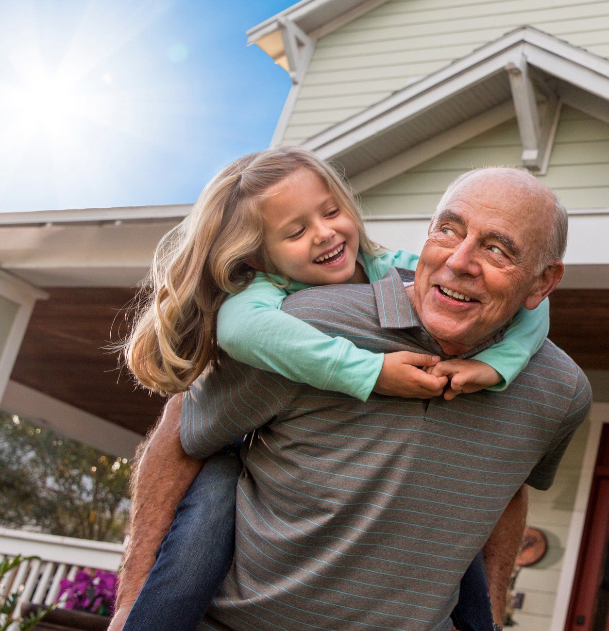 A man is carrying a little girl on his shoulders in front of a house.