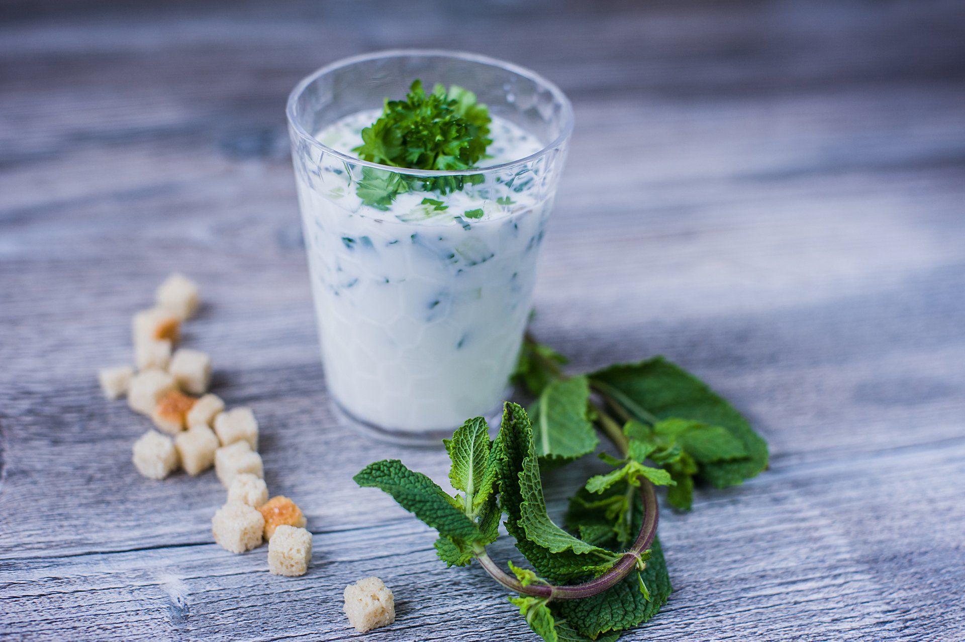 A glass of yogurt with mint leaves and croutons on a wooden table.