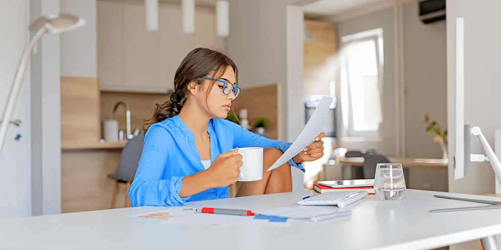 A woman is sitting at a table holding a cup of coffee and reading a piece of paper.