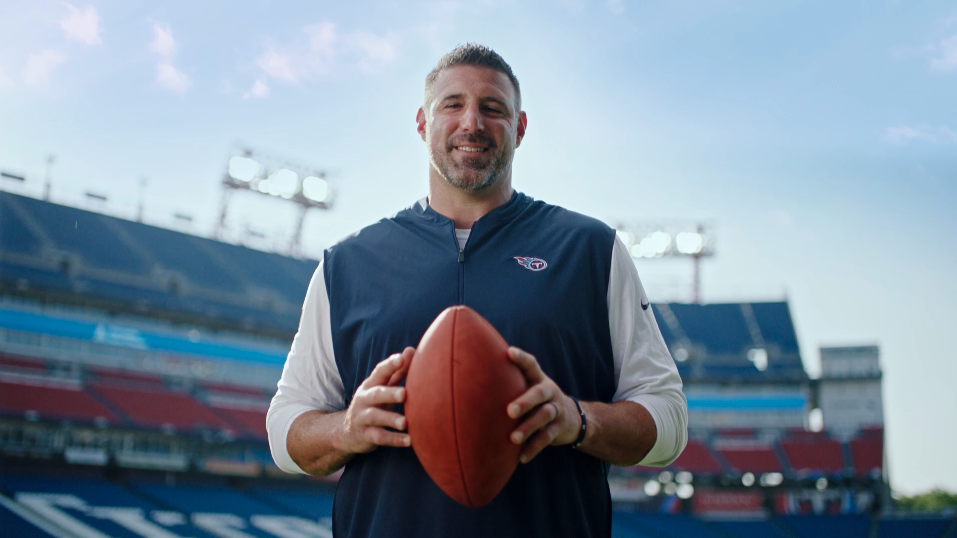 Mike Vrabel is holding a football in front of a stadium.