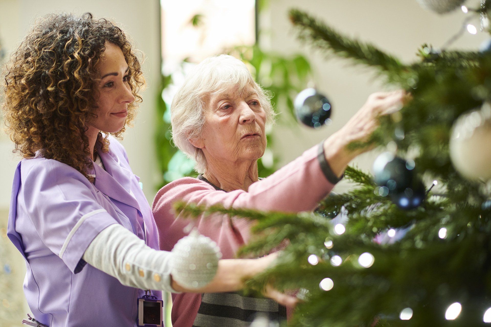 A nurse is helping an elderly woman decorate a christmas tree.