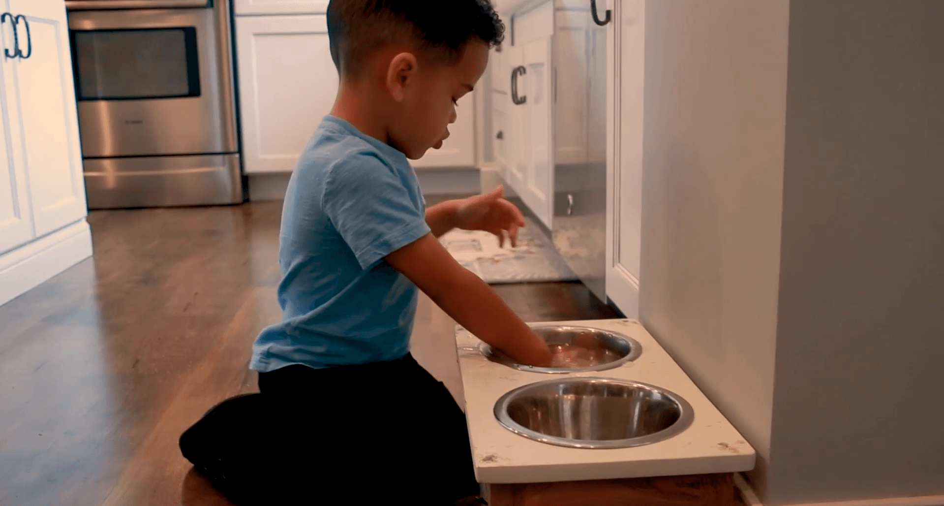 A young boy is kneeling on the floor in a kitchen playing with a dog bowl.