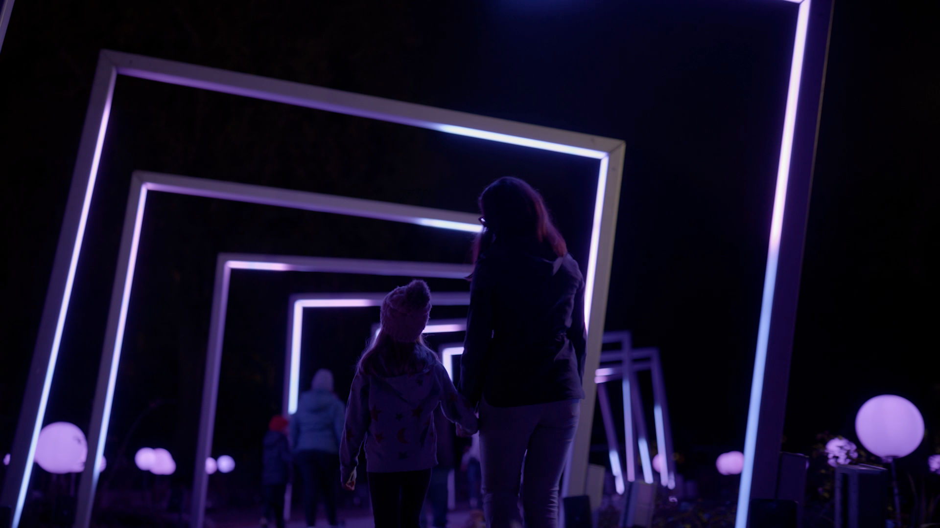 A woman and a child are walking through a tunnel of lights at night.