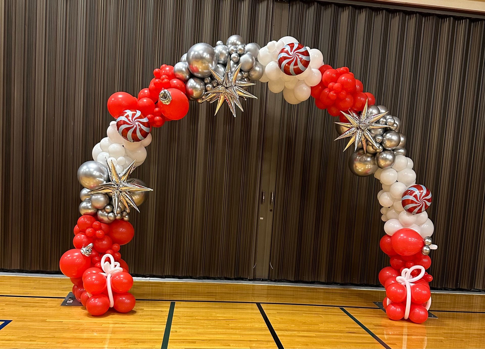 A red , white and silver balloon arch is sitting on a wooden floor.