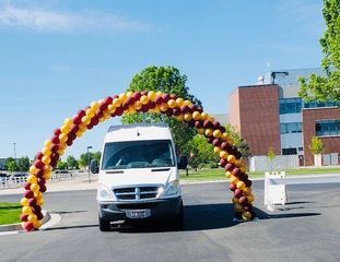 A white van is driving under a red and gold balloon arch