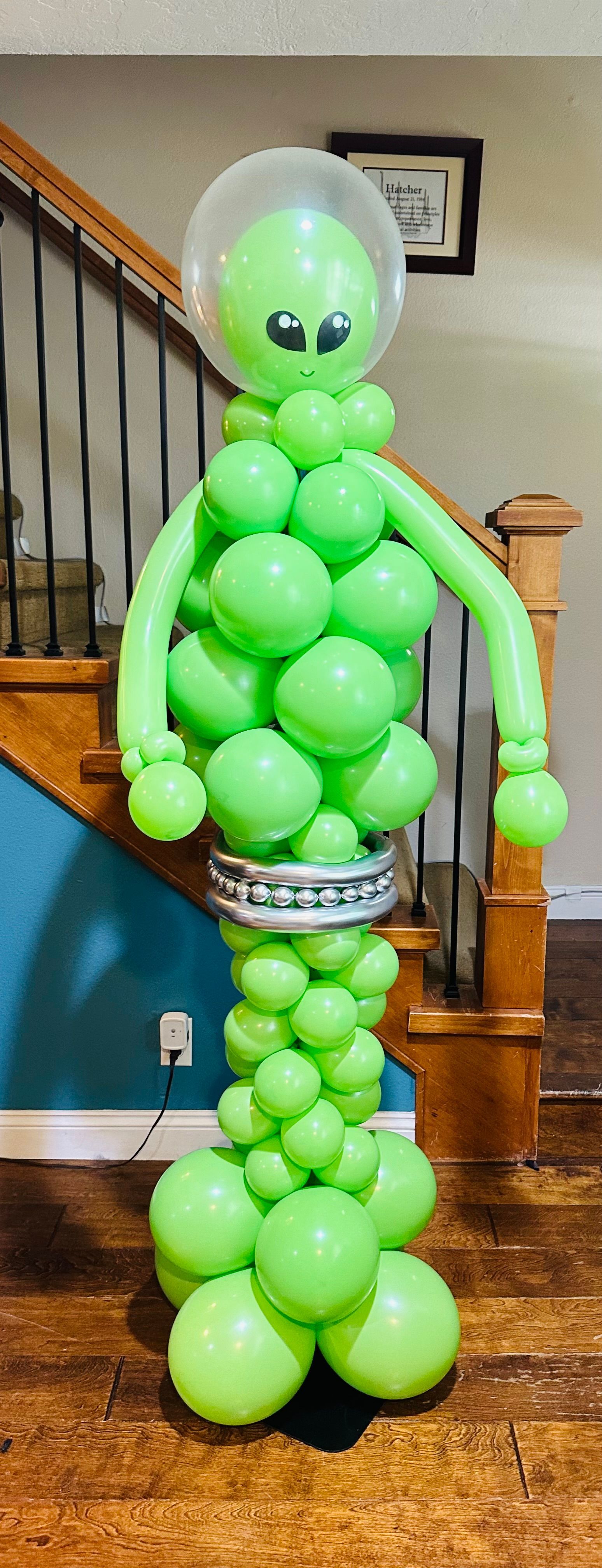 A green alien made out of balloons is standing next to a staircase.