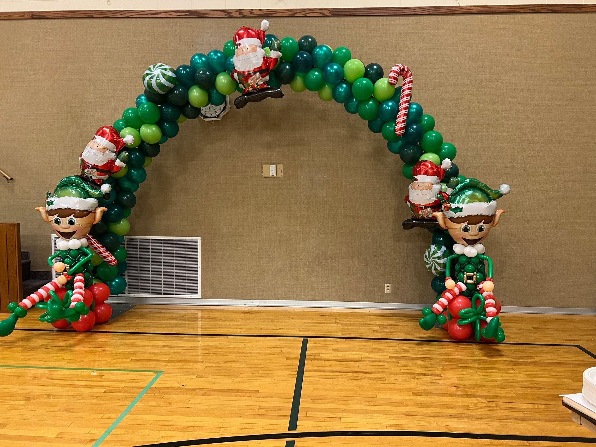 A balloon arch with santa claus and elves on it is in a gym.