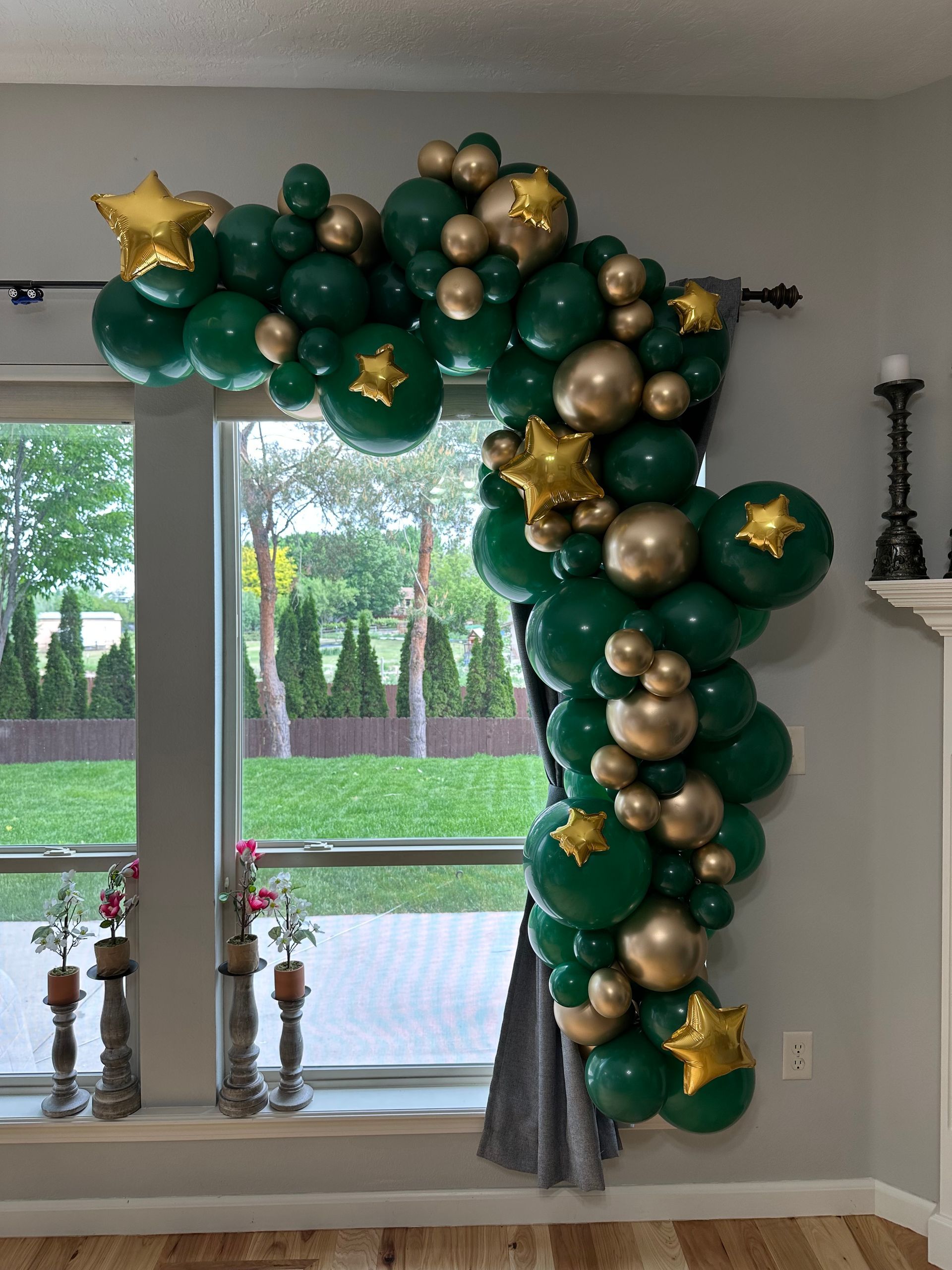 A green and gold balloon garland is hanging on a window in a living room.