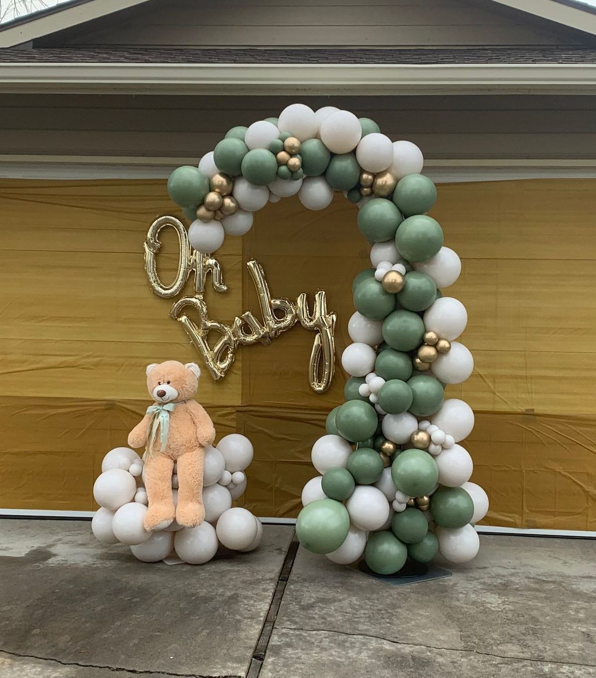A teddy bear is sitting in front of a balloon arch that says oh baby.