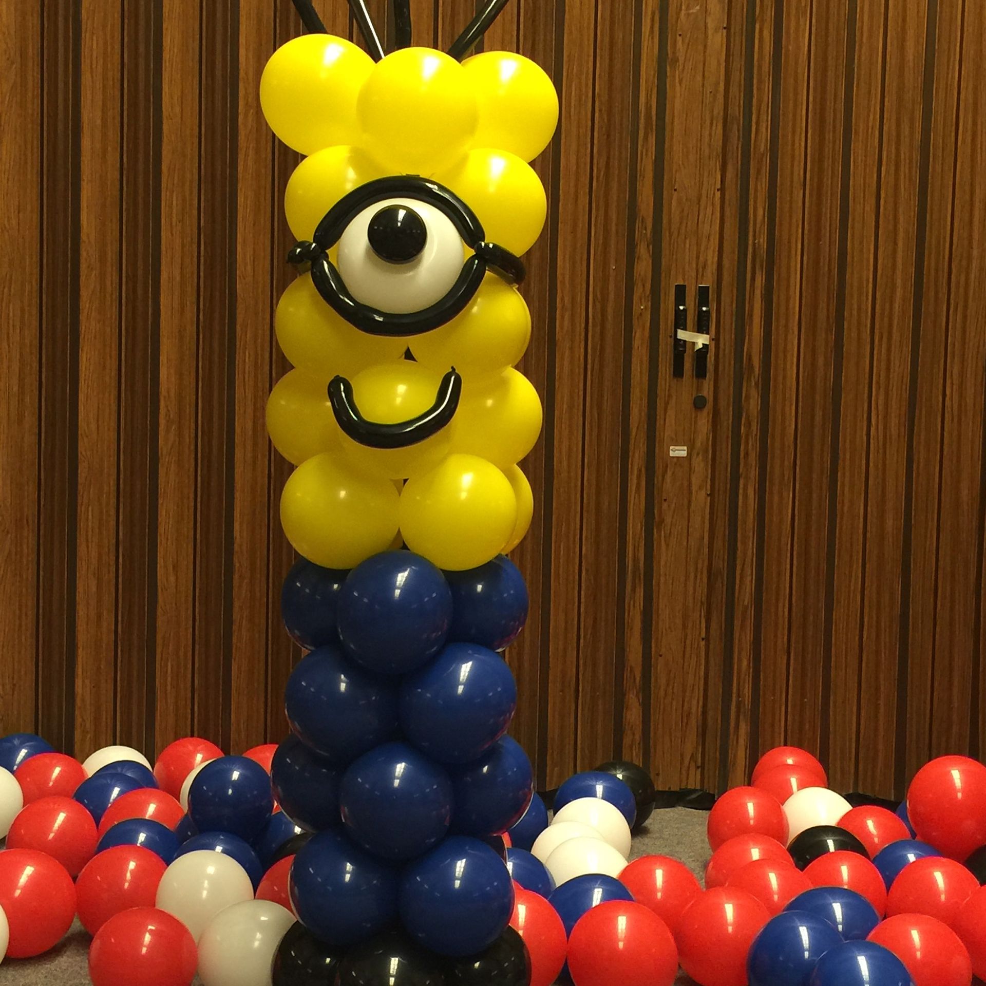 A balloon sculpture of a minion is surrounded by red white and blue balloons