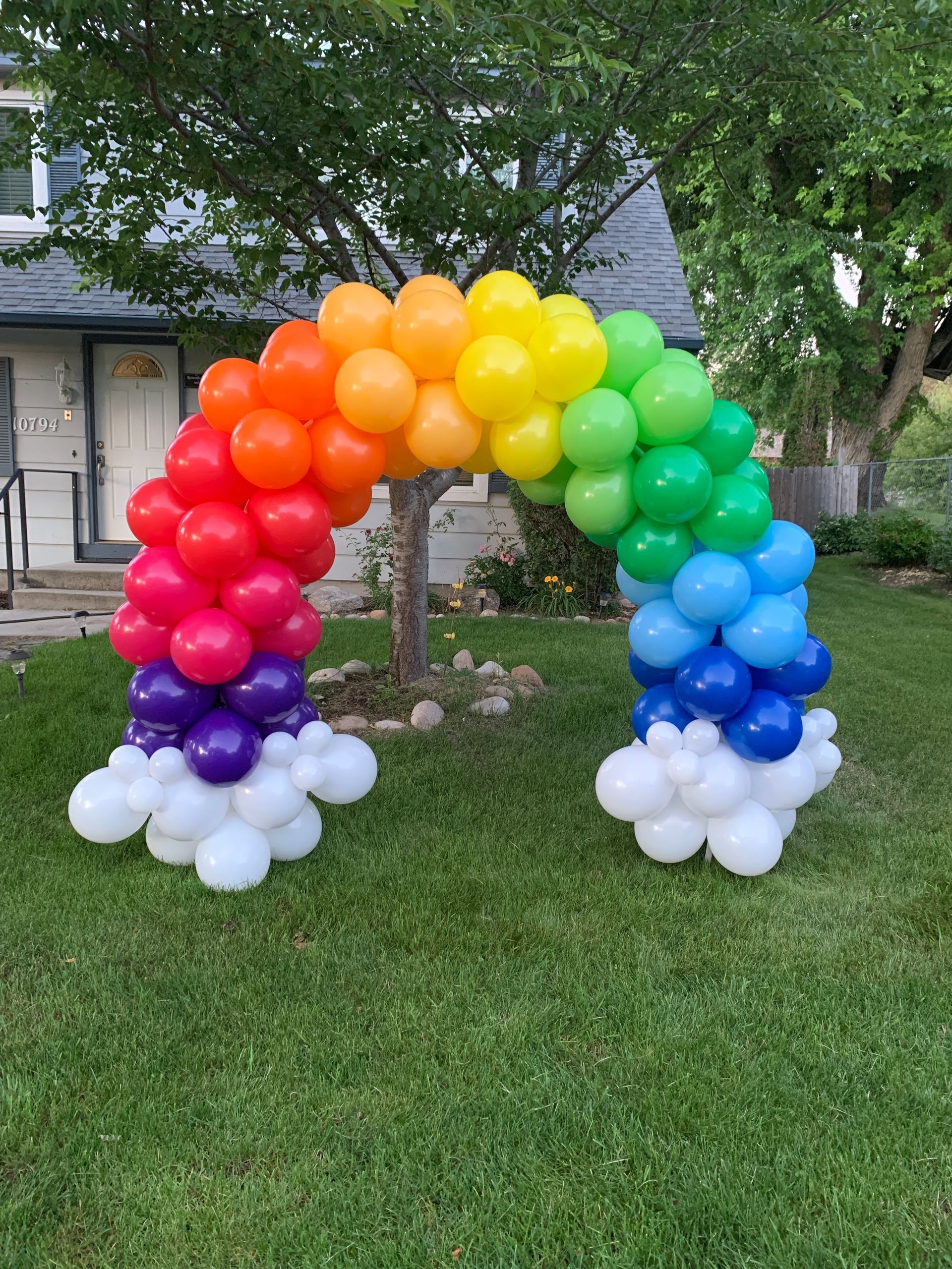 A rainbow balloon arch is sitting in the grass in front of a house.