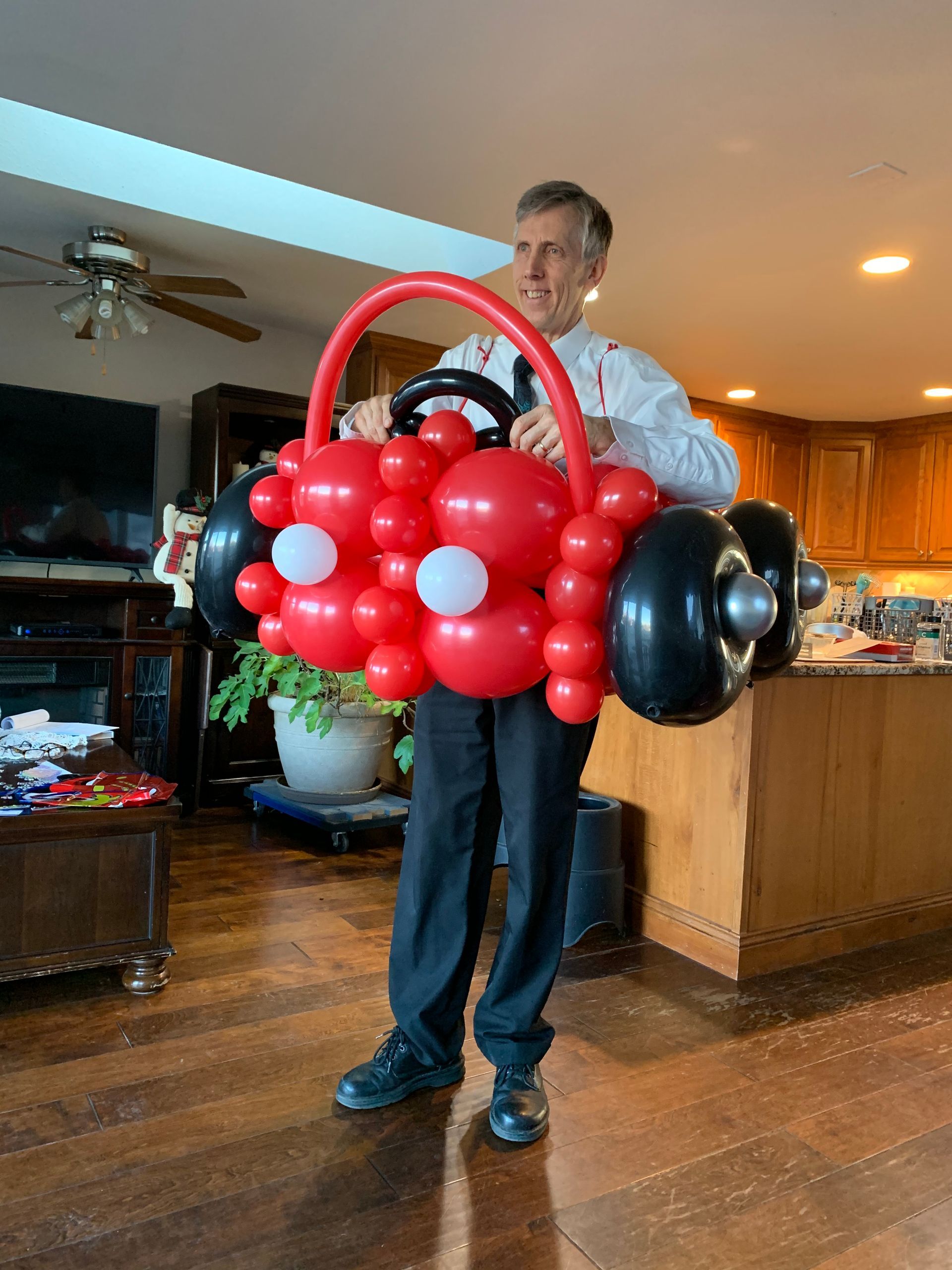 A man is holding a red car made of balloons in a living room.