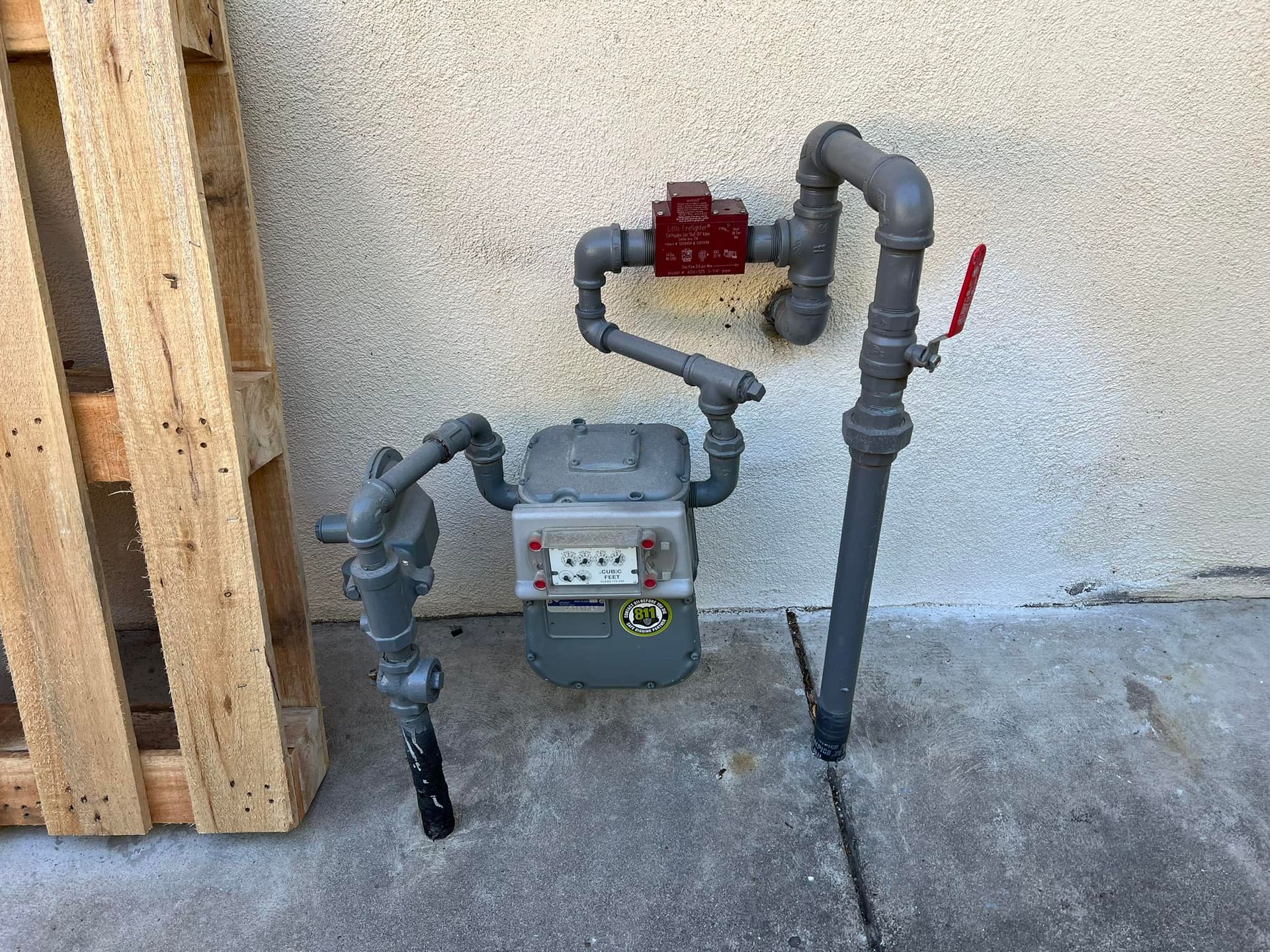 A gas meter is sitting on the sidewalk next to a wooden pallet.