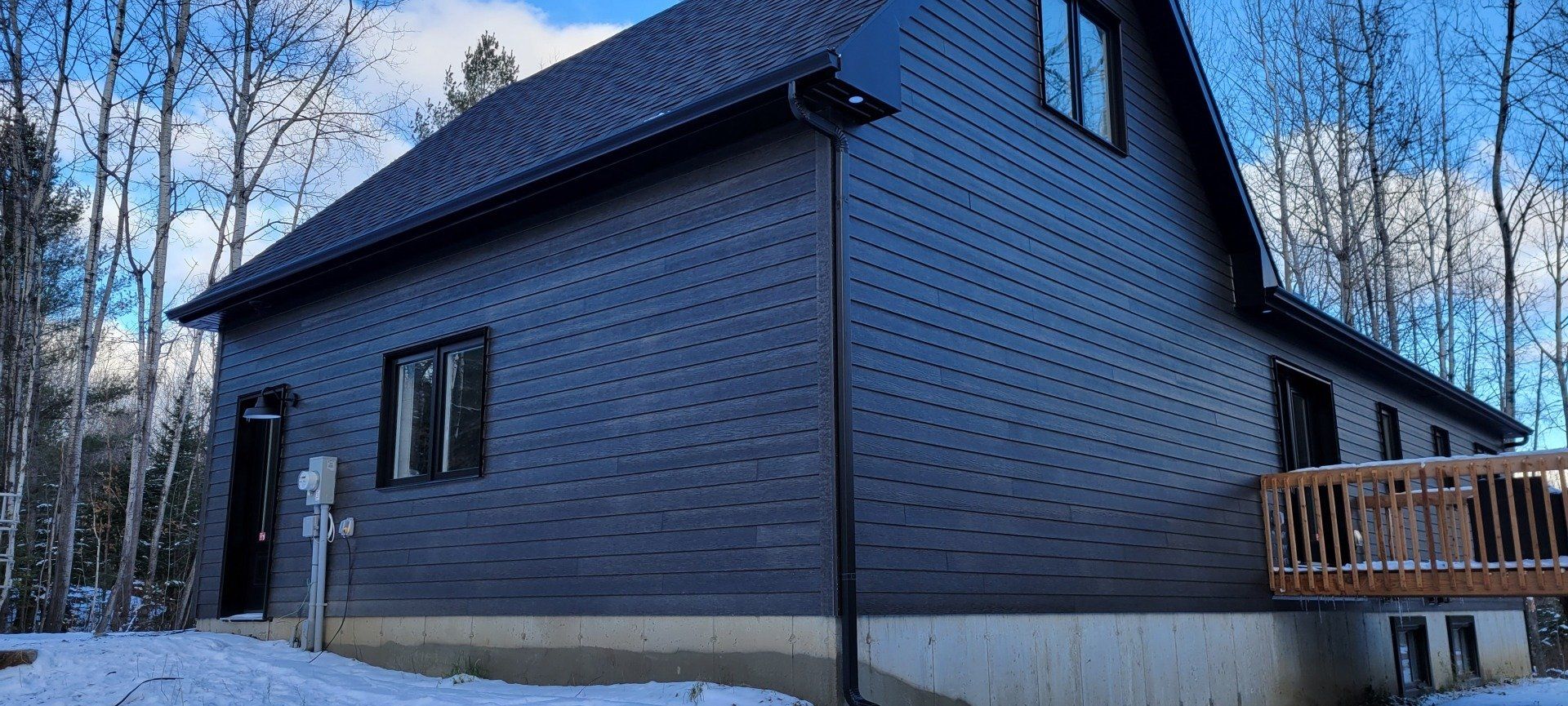 Une maison noire avec une terrasse en bois est entourée de neige et d'arbres.