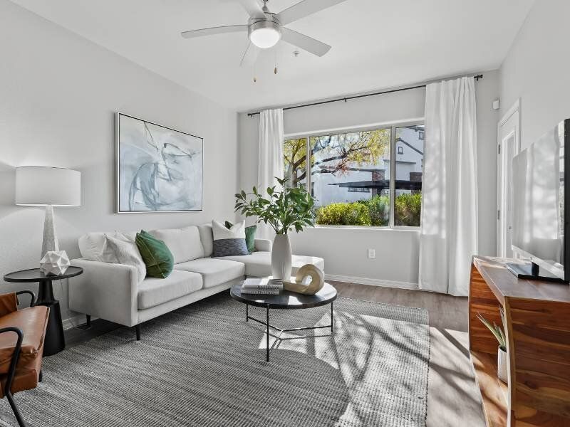 Bright living room with a white sofa, round glass coffee table, and large window with white curtains.