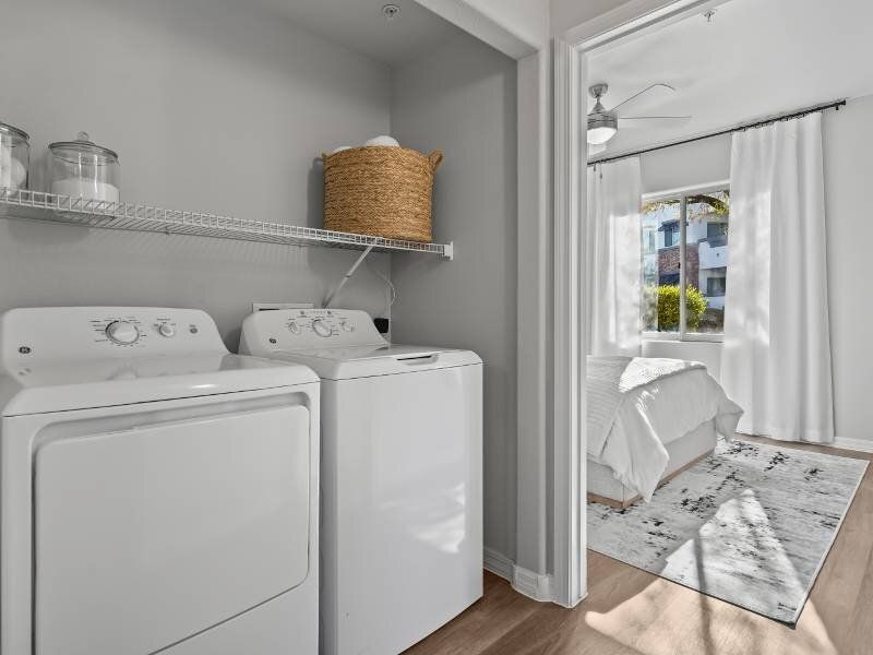 In-unit laundry closet with a washer and dryer, a wire shelf, and a basket; adjacent to a bright bedroom.