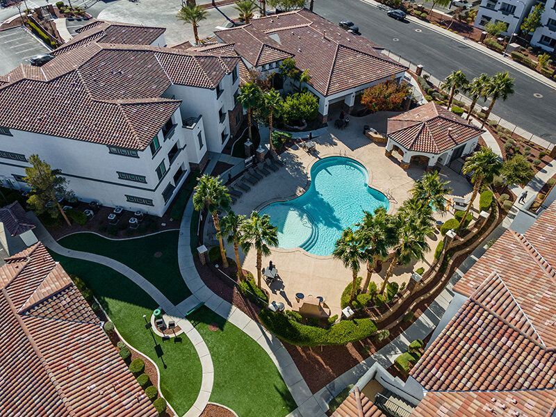 Aerial view of a resort-style apartment community with a curved pool, palm trees, and surrounding white buildings.