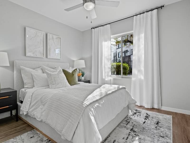 Modern bedroom in a light gray apartment with white bedding, ceiling fan, and a large window.