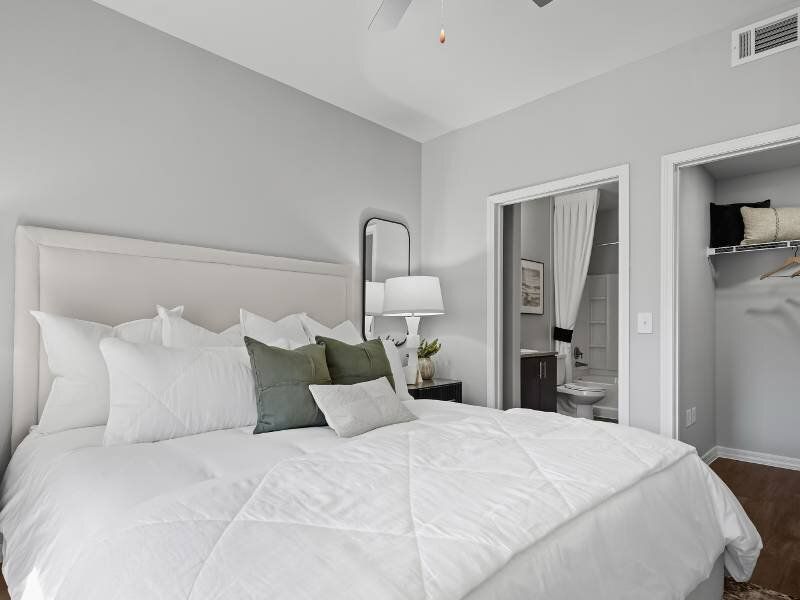 Modern bedroom with a large white bed, pillows, a nightstand with lamp, and an open closet beside the bathroom doorway.