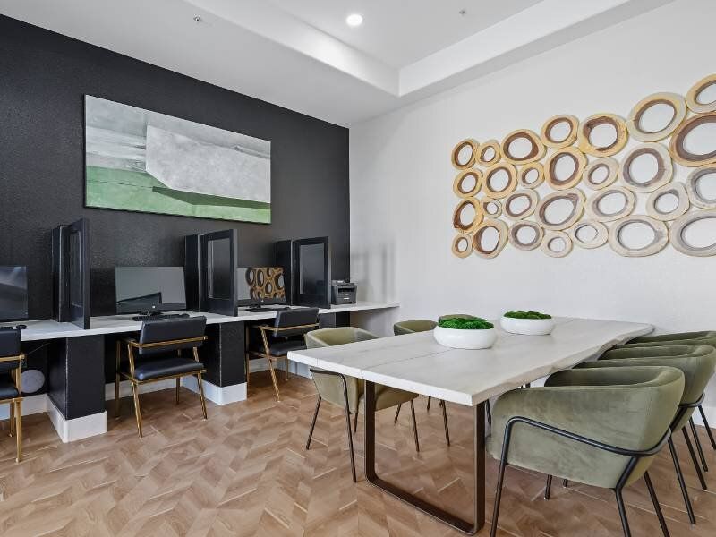 Modern community workspace with computer stations along a dark wall and a long white table with green chairs.