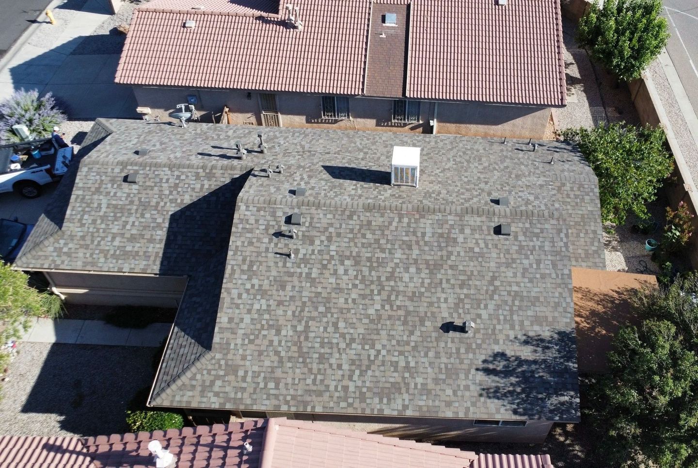 Overhead view of residential roofs with gray shingles and brown clay tile, with trees and a street in the background.