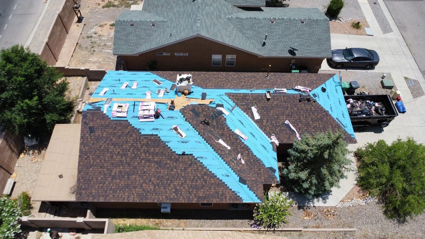 Roof of a house partially covered with blue underlayment, undergoing repair, with a dumpster nearby.