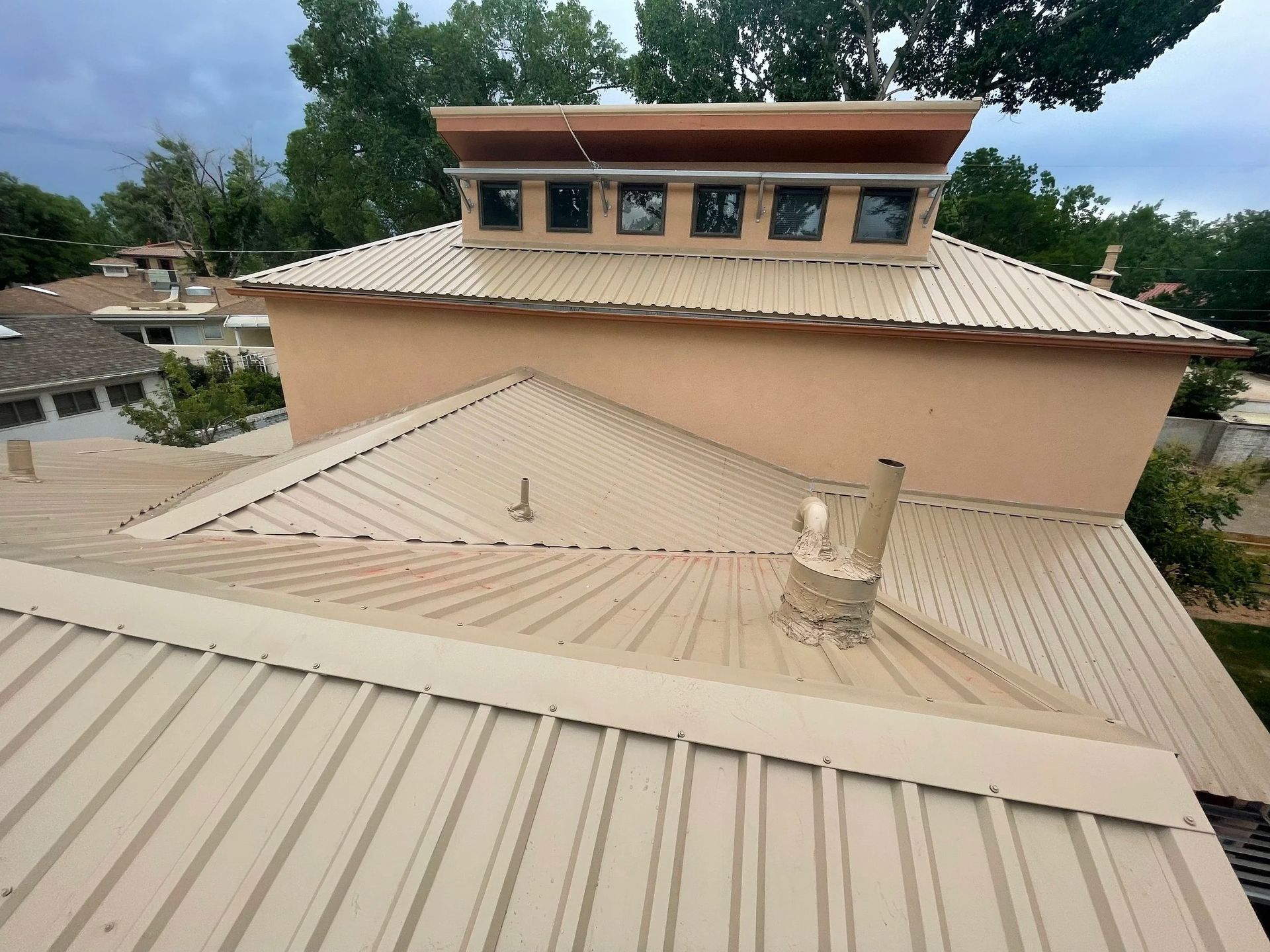 Tan metal roof with skylights and chimney against a cloudy sky, seen from a high angle.