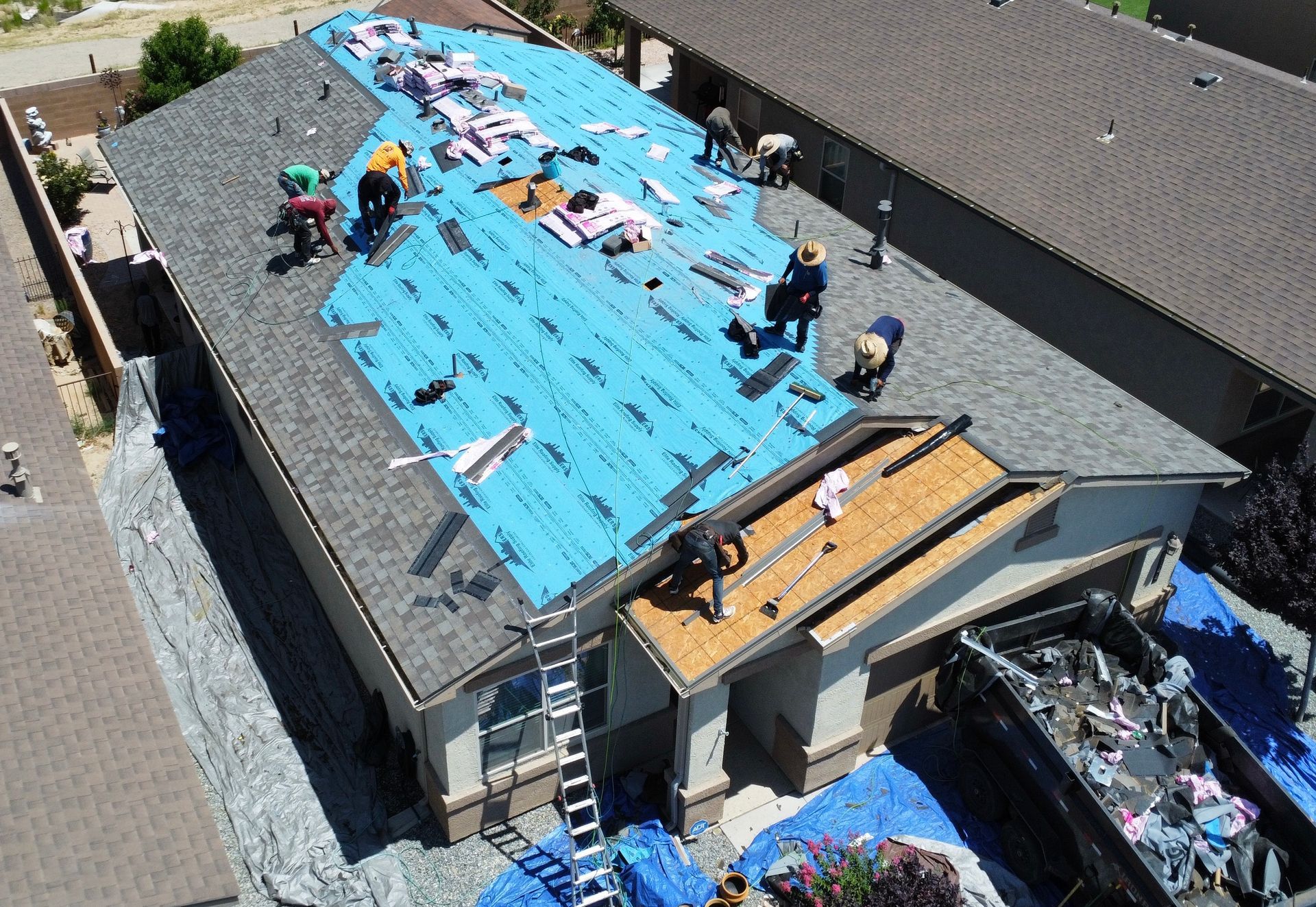 Roofers replacing shingles on a house; blue underlayment visible. Dumpster nearby.