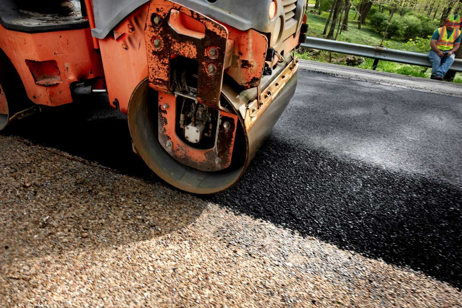 Steamroller paving fresh asphalt with worker in background.
