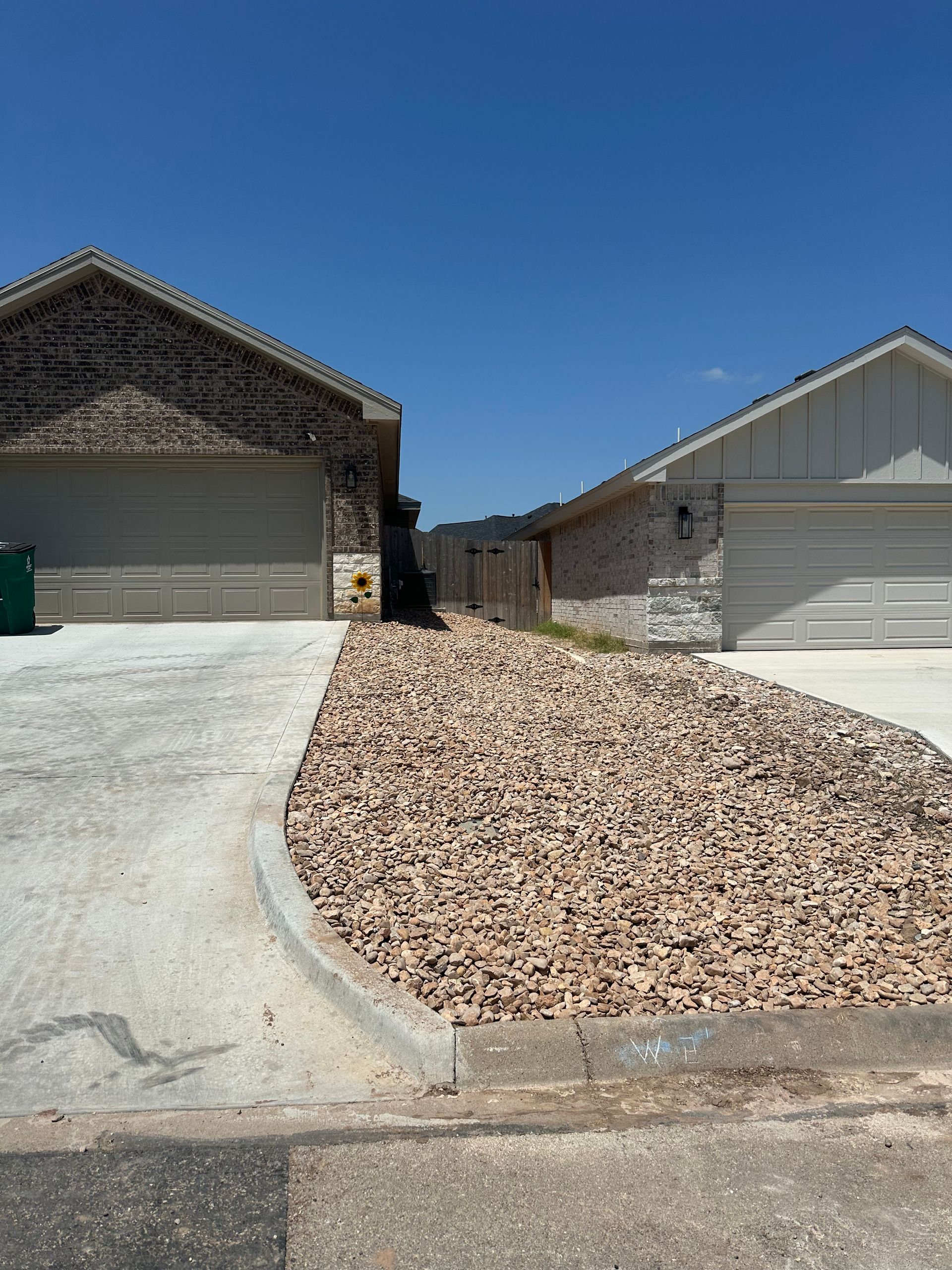 Two houses with driveways and a rock bed between them under a blue sky.