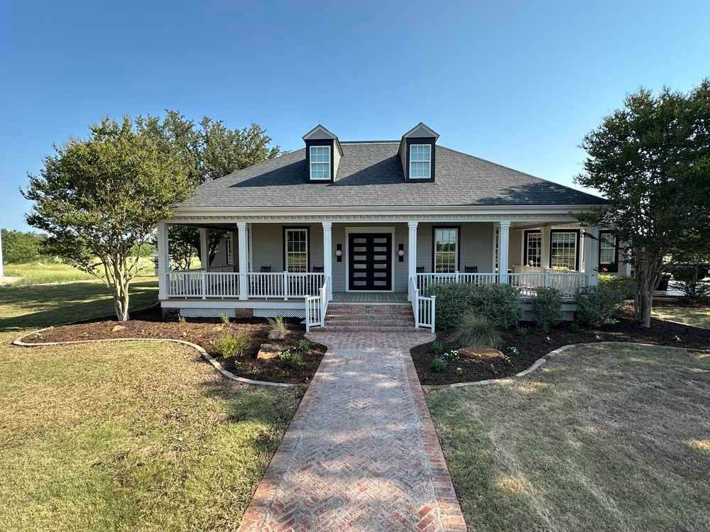 A single-story beige house with a brick pathway, a porch, and dormer windows, under a blue sky.