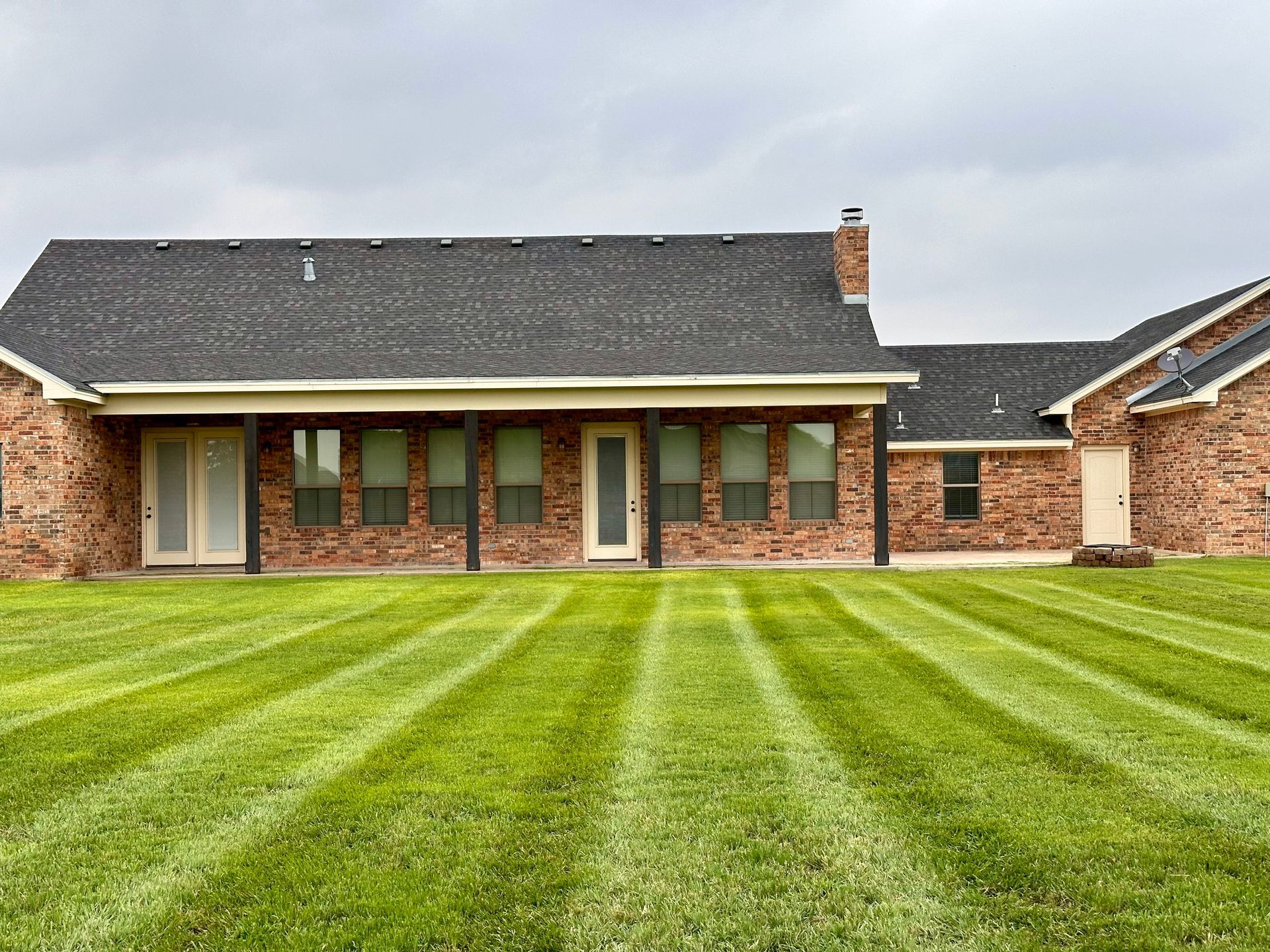 Rear view of a brick house with a green lawn striped from mowing, under a cloudy sky.