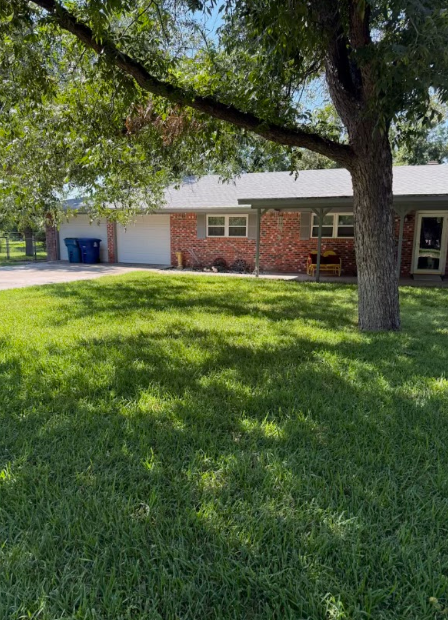 House with a brick facade, green lawn, and large tree casting a shadow.