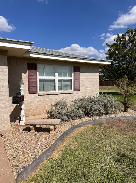 Brick house exterior with red shutters, small bench, and landscaped yard against blue sky.