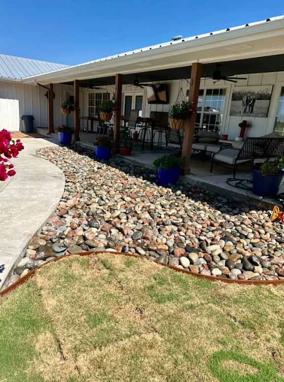 Rock garden border along a concrete walkway in front of a porch with blue pots and hanging plants; sunny outdoor setting.