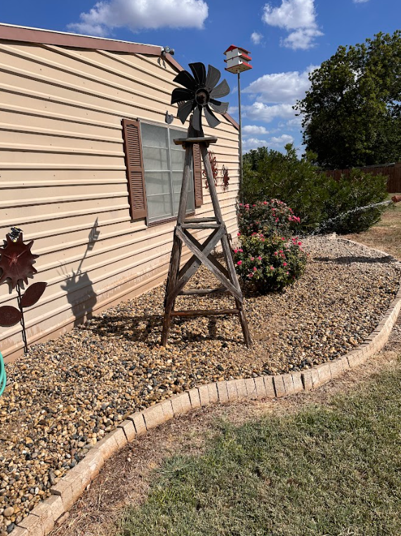 Windmill in a rock garden next to a house with a brown window and a birdhouse in the background.