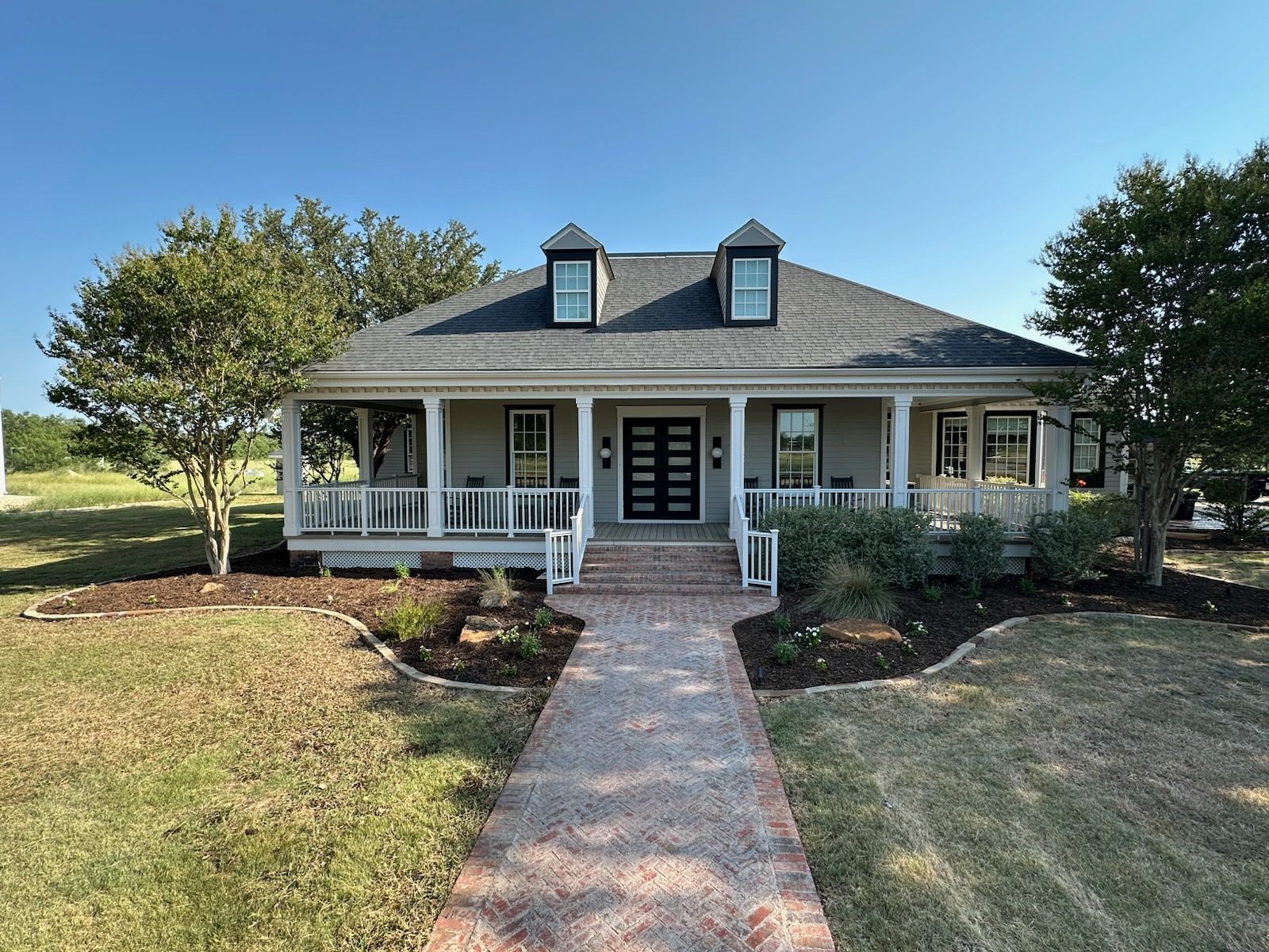 A one-story light gray house with a long porch, a brick walkway, and landscaping.