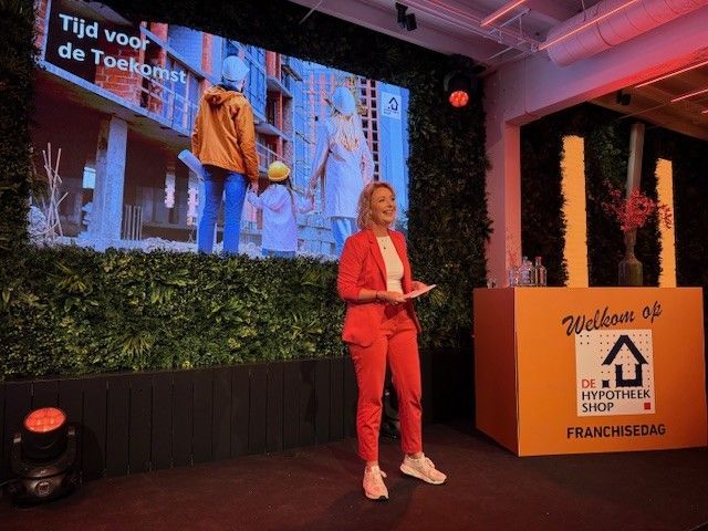 Vrouw in rood pak spreekt op een evenement met een scherm achter haar. Het logo van