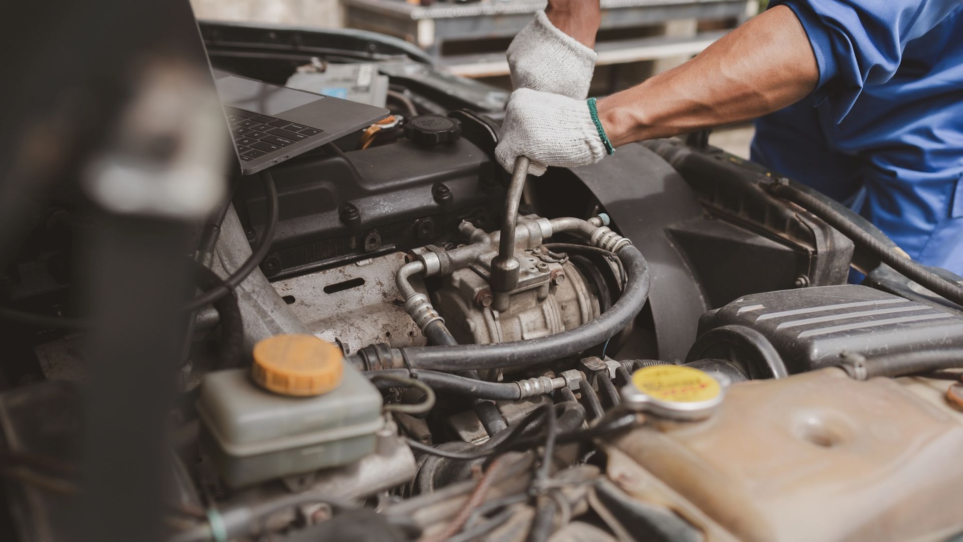 Mechanic in blue coveralls and gloves working on a car engine, using a wrench.
